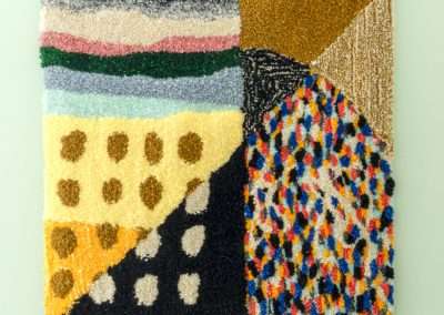 A textured, abstract rug featuring multiple colorful patterns such as stripes, polka dots, and speckled areas, with varied textures in shades of gold, black, blue, and green.