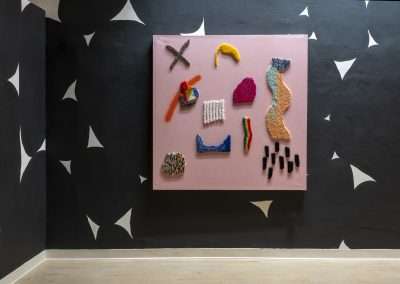 Modern art installation with a large pink canvas showcasing a variety of colorful, textured objects in abstract forms. Black wall background adorned with white star shapes.