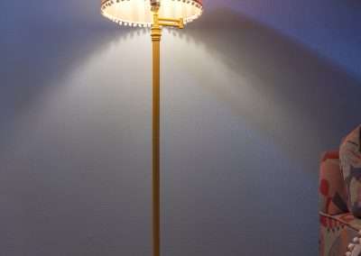 A floor lamp with a decorative shade illuminates a wall, casting a bright pattern of light in a dark room with a partial view of a floral-patterned chair to the right.