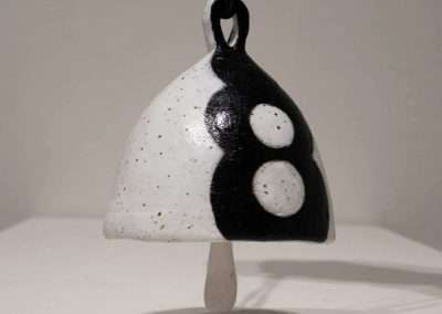 A ceramic bell suspended by a chain, featuring a black and white speckled pattern with three large black circles, casting a circular shadow on a light surface below. This piece is among my favorite things.