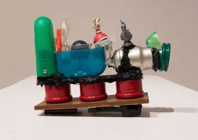 A mixed-media sculpture featuring various favorite things such as bottles and figurines assembled on a wooden base, connected with black material and mounted on red cylindrical supports.