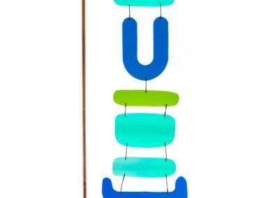 A colorful mobile sculpture, one of our favorite pieces, consisting of abstract blue and green shapes hanging on a wooden stand with a curved metal arm, set against a white background.