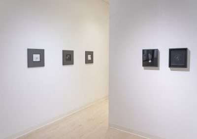 An art gallery interior showing a bright, clean space with white walls and wooden flooring. The walls display a series of small framed black and white photographs by Shoshannah White in a linear arrangement.