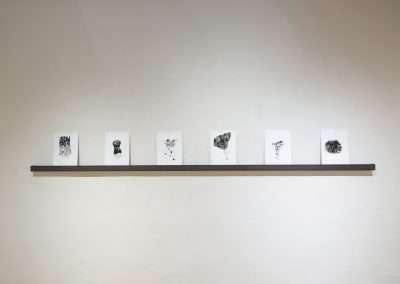 Six abstract artworks by Shoshannah White on white paper displayed in a row on a narrow shelf against a light-colored wall in a gallery.