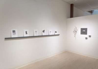 A modern art gallery room with a series of black and white abstract artworks by Shoshannah White displayed on a gray shelf and white walls. The room is brightly lit and has a wooden floor.