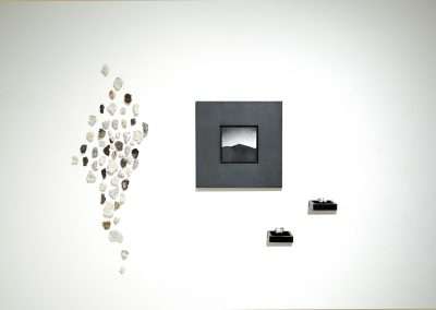 A modern art installation by Shoshannah White featuring a scattered arrangement of translucent rocks on a white wall, leading towards a black frame with a monochrome mountain landscape and two isolated small geometric sculptures.