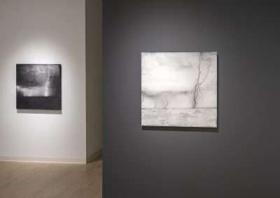 Two abstract paintings by Shoshannah White in a modern art gallery, one featuring darker tones on the left, the other with lighter tones and a prominent crack pattern on the right, displayed on a gray wall.