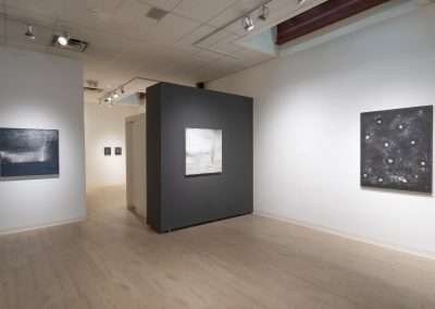 A modern art gallery featuring several abstract paintings on white walls includes a dark gray partition in the center displaying a large Shoshannah White painting. The floor is light wood, and the ceiling has light fixtures illuminating the artworks.
