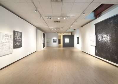 Spacious, well-lit art gallery interior with numerous diverse paintings and photographs by Shoshannah White displayed on white walls, featuring a polished wooden floor.