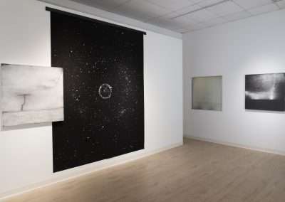 Art gallery interior featuring three abstract artworks: a large cosmic-themed canvas by Shoshannah White in the center, a cracked surface canvas on the left, and a soft-toned blurry image on the right.