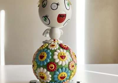 A colorful, decorative Takashi Murakami figurine with a floral-patterned body and a face featuring whimsical, cartoonish features, standing on a white surface surrounded by Murakami flower pins.