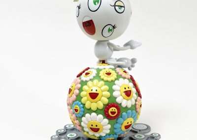 A whimsical figurine with a large head, wide eyes, and an open mouth sits atop a colorful sphere covered in smiling flowers. The sphere rests on a flat surface adorned with circular patterns. The background is plain white.