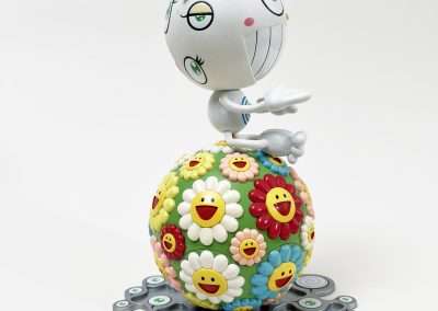 A whimsical, white character with large green eyes and a wide, toothy grin sits atop a colorful, spherical object covered in cartoonish flowers with smiley faces. The base includes multiple round shapes adorned with a single green eye each.
