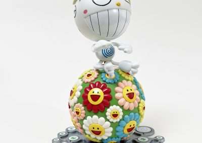 A whimsical figurine featuring a character with large eyes, a wide, toothy smile, and spiky hair, sitting atop a colorful sphere covered with smiling, cartoonish flowers. The sphere is surrounded by small, flat, round shapes with eyes.
