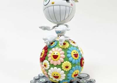 A whimsical figurine featuring a winking character with a large, spherical head sitting on top of a multicolored ball adorned with smiling flower faces. Below the ball are scattered smaller, smiling eye-like designs. The overall aesthetic is playful and colorful.