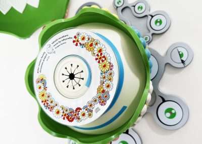 A top view of a colorful flower-shaped toy with a floral pattern around its circular center. Surrounding it are several gray, circular pieces with green eye-like designs. The toy seems to be disassembled and is laid out on a white surface.
