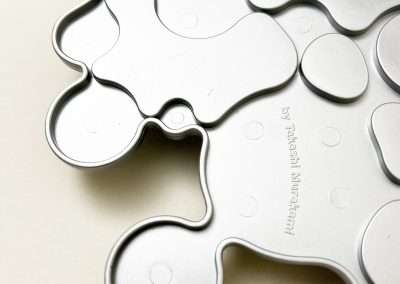 Close-up of a seamless, interlocking metal trivet with an abstract, organic shape resembling puzzle pieces or molecules. The surface has several raised dots, and some text is faintly visible on one piece. The background is a plain, light-colored surface.