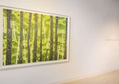 A framed painting, reminiscent of Alex Katz's style, depicts several trees with green foliage and light filtering through the leaves. It hangs on a white wall that is otherwise bare except for faint text in the background that reads, "Happy Birthday, Aya Kato.