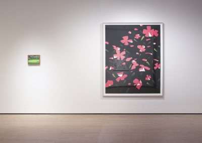 A minimalist gallery space features two framed artworks on a white wall. On the left is a small, detailed landscape painting, and on the right is a large abstract piece with pink flowers on a dark background. The floor is light wood.