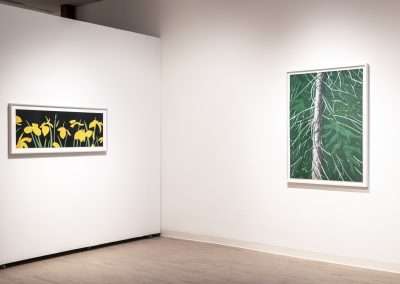 A gallery wall featuring two framed artworks: the left one is a horizontal painting of yellow flowers with green stems against a black background, and the right one is a vertical painting depicting the green leaves and trunk of a tree.