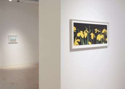 A minimalist art gallery features two framed paintings. The foreground shows a rectangular painting of yellow flowers against a black background on a white wall. In the background, there is a smaller abstract painting with light colors displayed on a wall.