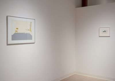 A minimalist art gallery wall displays two framed paintings. The left painting features abstract shapes in muted colors; the right painting is smaller, featuring a delicate, understated composition. The white walls create a clean, serene setting.