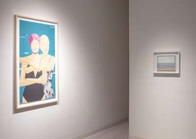 A minimalist art gallery features two framed artworks on white walls. The left frame displays a modern, colorful painting of two abstract human figures in swimwear. The right frame shows a smaller, subdued landscape painting of a beach scene.