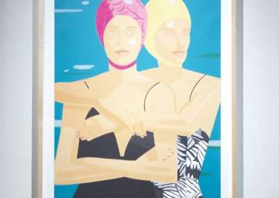 A framed painting of two individuals wearing swimming caps and swimsuits, standing in water. The person on the left has a pink cap and a black swimsuit, and the person on the right has a yellow cap and a black-and-white patterned swimsuit, with their arms around each other.