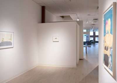 A minimalistic art gallery with white walls and light wood flooring features three framed artworks. Two paintings are on the left wall, one near the front and one in the back. A large, colorful piece is displayed prominently on the right wall near the entrance.