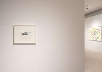 A minimalistic art gallery with white walls displaying framed artworks. A small painting of a house is on the left wall, while larger, colorful floral paintings can be glimpsed around the corner on the right wall. The gallery space is well-lit and uncluttered.