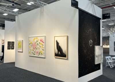 A gallery display features various artworks: stylized daisies, abstract squares, a monochromatic figure, an intricate starry sky, and other mixed media pieces on paper. The white walls and bright lighting enhance the art in the modern exhibit space.