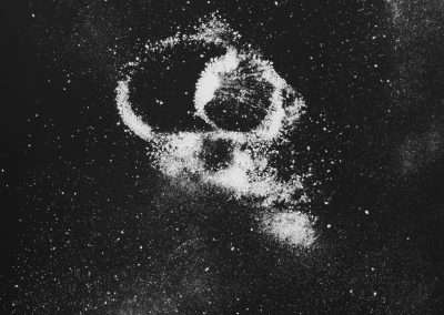 An abstract image of white salt or powder arranged loosely in a rough spiral shape against a black background, creating a cosmic or galaxy-like appearance. Fine particles are scattered around the central formation, adding a sense of depth and texture.