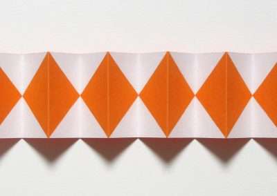 A paper strip featuring a zigzag fold pattern with alternating orange and white diamond shapes is displayed against a white background. The geometric design creates a visually striking effect, casting shadows beneath the ridges of the folds.