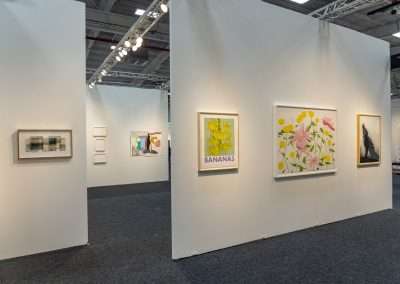 A contemporary art gallery features several framed artworks hanging on white partition walls. Among the visible pieces are a floral painting on paper, an artwork labeled "BANANAS" with yellow bananas, and a few abstract and minimalist works. The space is well-lit, showcasing the diverse art beautifully.