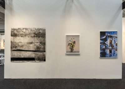 A gallery wall displaying three framed artworks: from left to right, a large black and white abstract piece, a colorful still life featuring a vase of flowers, and a vibrant photograph of boats reflected in water. The floor is dark carpeted, and two white chairs are partially visible amidst this curated paper art collection.