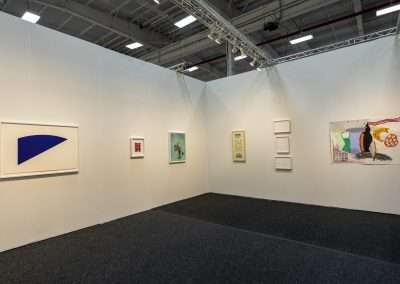 An art gallery features a corner setup displaying minimalist and abstract artwork on paper. The walls have various framed pieces, including a blue shape, a grid pattern, and colorful, intricate designs. The well-lit space highlights each piece of Art on Paper clearly.