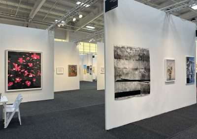 A contemporary art gallery with white walls exhibiting various artworks, including Art on Paper. Paintings of different styles and sizes are displayed, featuring a large one with pink flowers to the left and an abstract black-and-white piece to the right. A white chair and table are in the foreground.