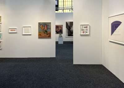 An art gallery featuring various framed artworks on white walls. The pieces range from abstract designs to detailed paintings, with some exquisite art on paper. In the center of the gallery is an open space leading to another section with more artwork. The floor is carpeted in dark gray.
