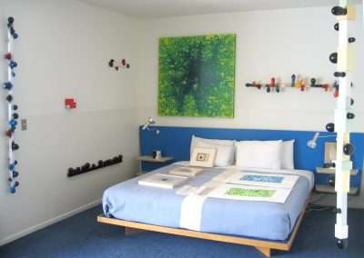 A modern bedroom features a blue bed with art pieces on it, surrounded by white and light blue walls. There are colorful, abstract wall sculptures and a large green abstract painting above the bed. The room has contemporary decor with a mix of geometric and organic forms.