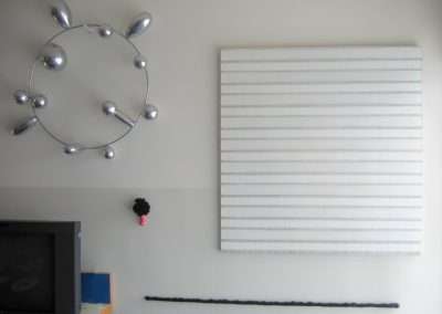 A minimalist room featuring a TV in the bottom left corner, a modern metallic wall sculpture resembling a molecule on the left wall, a large striped white canvas hung centrally, a small black flower decoration, and a long black rod displayed horizontally.