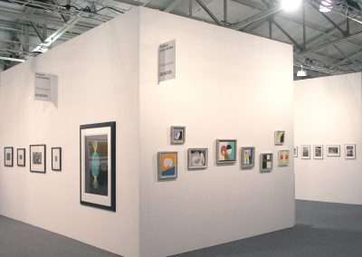 A white-walled art gallery displaying various framed artworks of different sizes. The pieces include abstract and modern art. The gallery is well-lit with overhead lights and has a clean, minimalist design. Labels on the walls indicate gallery sections.