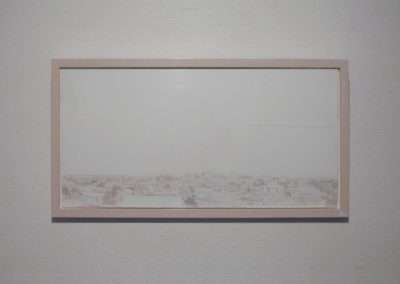 A rectangular frame with a light wooden border hangs on a plain white wall. The framed content appears to display a faint, barely visible landscape or cityscape, characterized by extremely light, almost indistinct, details and colors.
