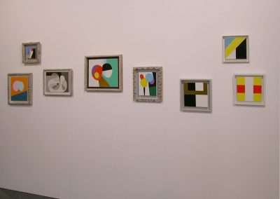 An art gallery wall displays seven framed abstract paintings, each with vibrant geometric shapes and contrasting colors. The artworks vary in size and frame style, creating an eclectic and visually engaging presentation.