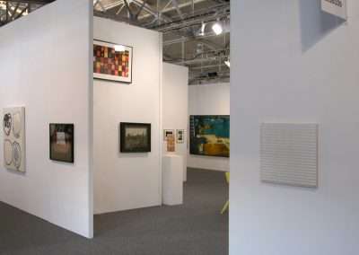 Art gallery space with white walls displaying various framed artworks. There is a mixture of abstract, landscape, and modern art pieces. The ceiling has exposed architectural elements and bright overhead lighting. The floor is carpeted in dark gray.