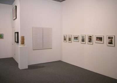 A minimalistic art gallery features various pieces. The left wall holds a series of artworks including a framed piece and a small sculpture on a pedestal. The far right wall displays a horizontal line of framed photographs. The floor is carpeted.