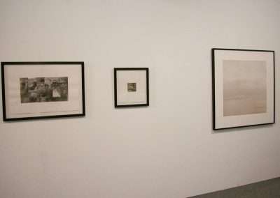 A white wall displays three framed artworks in a minimalist gallery setting. Arranged in a horizontal line, the frames vary in size, with the largest on the right, a medium-sized piece on the left, and a small one in the middle. The floor is carpeted.