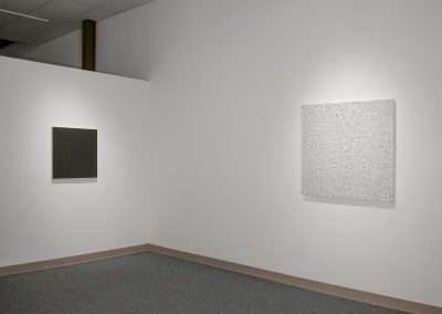A minimalist gallery room features two square artworks mounted on a white wall. The left piece is a solid dark-colored square, while the right piece, reminiscent of Teo Gonzalez's style, is a light-colored square with intricate, abstract patterns. The room is well-lit and has a gray carpeted floor.
