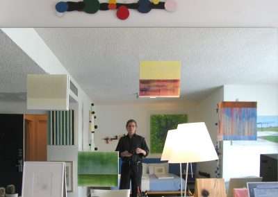 A person stands in front of a mirror in an art-filled room, holding a camera. Various colorful paintings and decorations hang around the space. On a shelf below the mirror, there are art supplies, a small potted cactus, and a computer screen is partially visible.