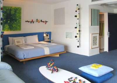 A modern, brightly lit bedroom features a blue bed with a white stripe in the center, a low white coffee table with colorful geometric toys, and a blue couch. The room has abstract wall art, a blue carpet, and decorative elements near the entrance.