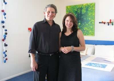 A smiling man and woman stand side by side in a brightly lit room. The man wears a dark shirt and trousers, while the woman is dressed in a black dress. Behind them is a bed with colorful throws and a green abstract painting on the wall.